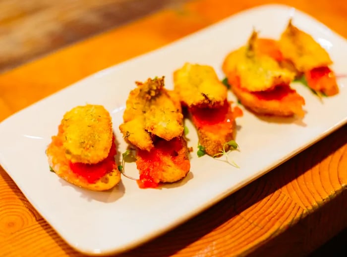 Five small slices of bread topped with a mixture of tomato and large fried sardines arranged on a long plate.