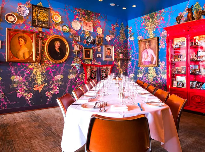 A long dinner table adorned with a white tablecloth, complemented by leather-backed chairs. The blue-painted walls are decorated with an array of eclectic artwork in ornate frames.