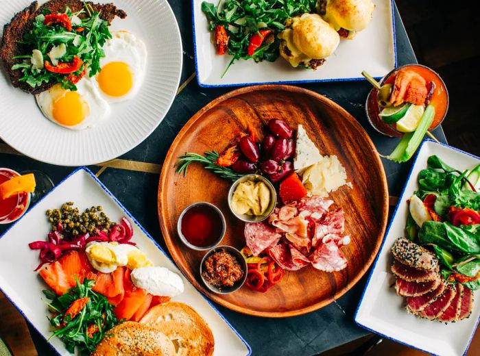 An overhead view of a table adorned with a variety of dishes, including a crudité and prosciutto platter, seared tuna, steak and eggs, and a brunch assortment.