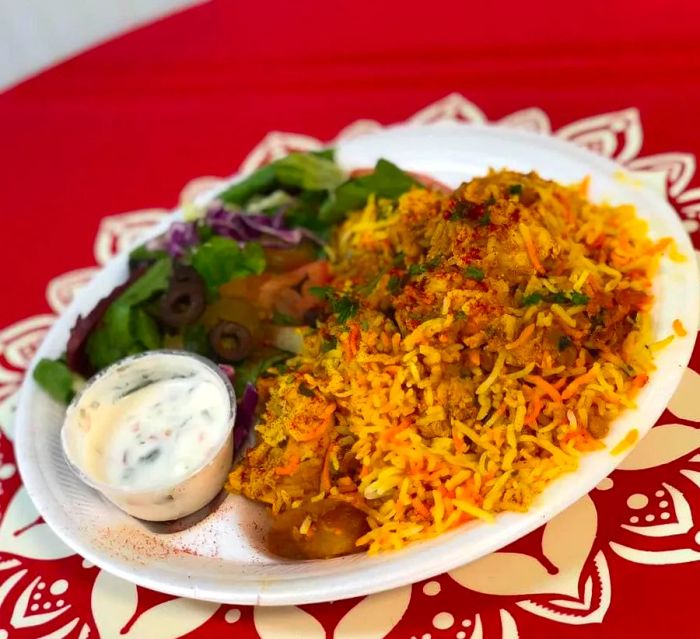 A vibrant plate of biryani adorned with pieces of meat, accompanied by a cup of white sauce and a side salad.