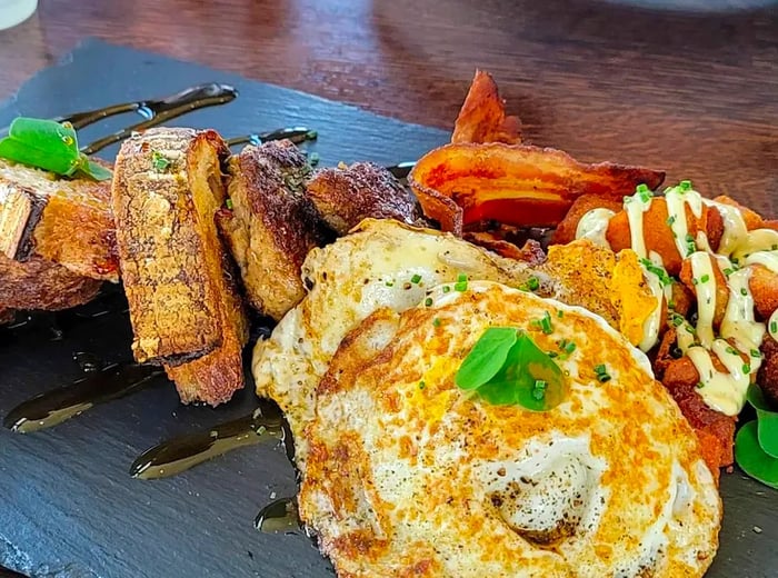 A slate board featuring eggs, toast, patatas bravas drizzled with white sauce, and crispy bacon.