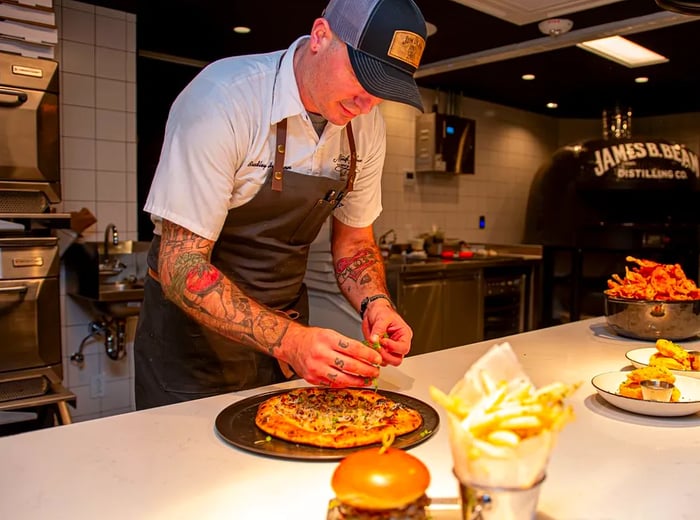 A chef skillfully prepares a thick flatbread on a kitchen counter, surrounded by various dishes in different stages of preparation. In the background, a large pizza oven proudly displays the James B. Beam Distilling Co. logo.