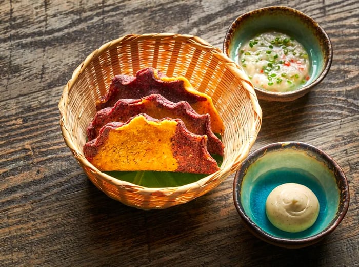 A basket containing three yellow and blue empanadas, accompanied by a small bowl of crab-shrimp dip and a dish of aioli.