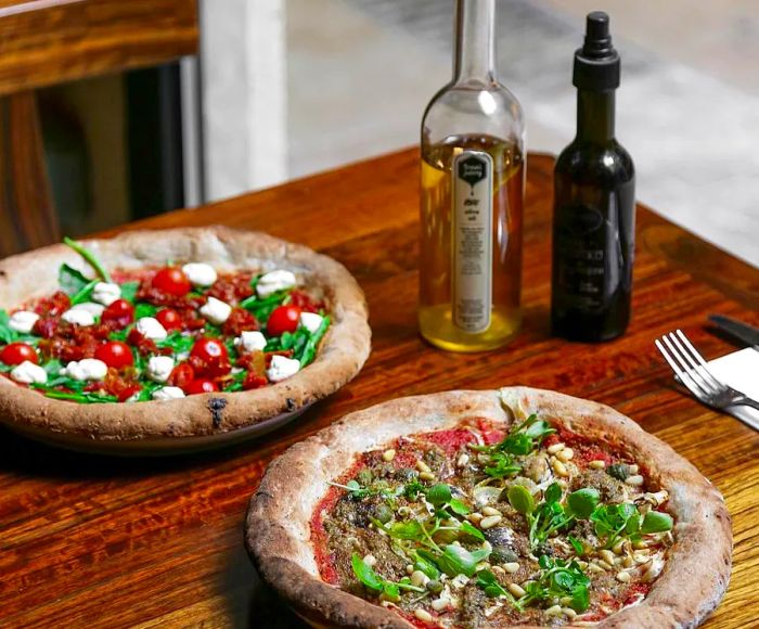 A pair of vegetable-packed pizzas on a beautiful wooden table accompanied by a bottle of olive oil.