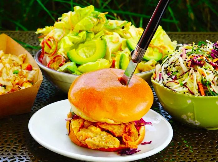 A fried chicken sandwich perched on a fluffy bun, secured with a steak knife, accompanied by bowls of side dishes.