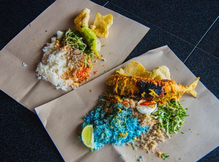 Fried fish and vegetables served on wax paper alongside vibrant blue rice, white rice, and various accompaniments.