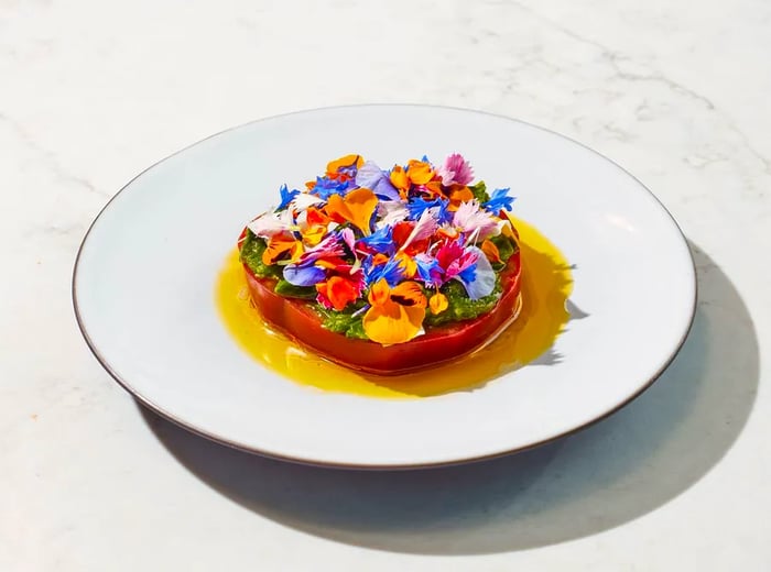 A slice of tomato adorned with a colorful array of flowers in a pool of golden sauce.