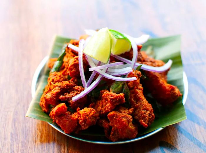 A serving of fried chicken pieces arranged on a leaf, garnished with sliced red onion and lime wedges.