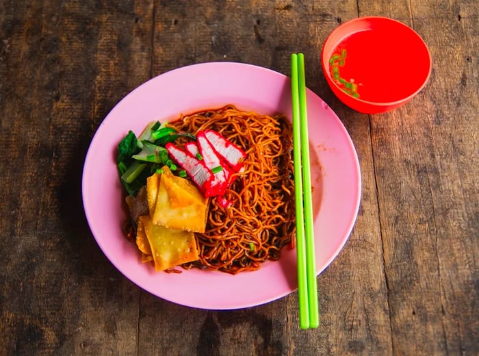 A bowl containing delicate noodles topped with wontons, vibrant red pork slices, greens, and sauce.