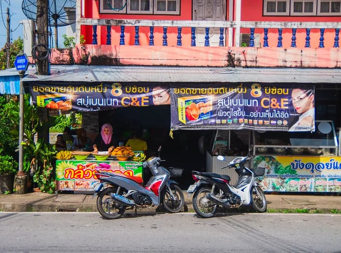 A woman in a vivid pink hijab prepares her roadside stall under an awning adorned with various advertisements in Thai.