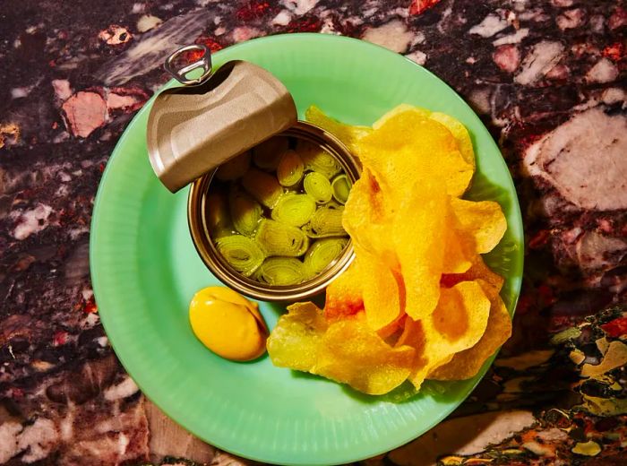 An overhead view of a tin filled with preserved leeks, accompanied by chips.