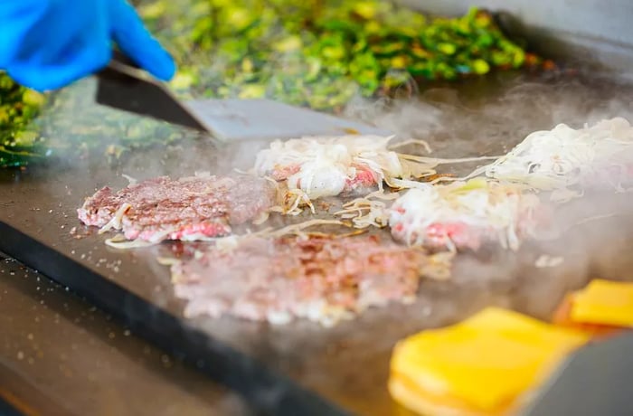 Burger patties pressed onto the griddle with onions as a cook stands nearby with a spatula.