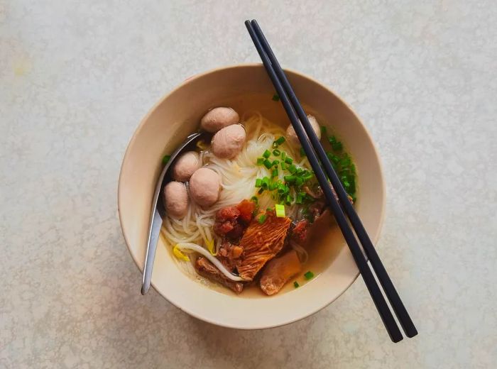 An aerial view of a bowl filled with broth, fish balls, slices of meat, and fresh herbs garnishing the dish.