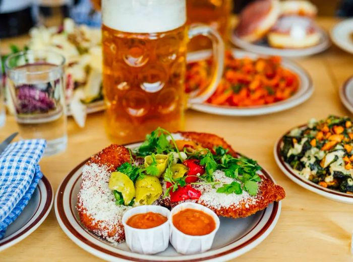 A large, thin schnitzel adorned with peppers and greens, accompanied by sauces next to a stein of beer.