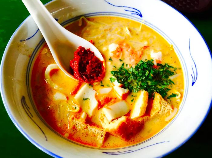 A bowl of Katong Laksa, a beloved spicy noodle soup, is displayed. This dish is a staple of Peranakan cuisine.