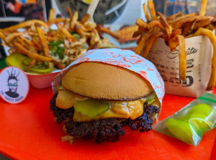 A smash burger topped with cheese and pickles, served on a tray with two portions of fries featuring various toppings.