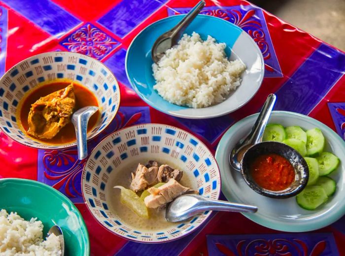 An array of dishes featuring chicken broth, rice, fish collar, and cucumbers served with dipping sauce, arranged on a vibrant tablecloth.