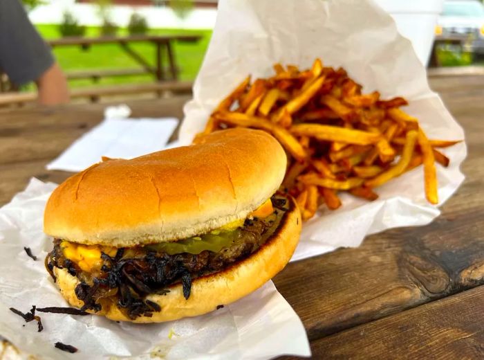 A burger topped with deeply charred onions rests on a picnic table next to a heap of fries.