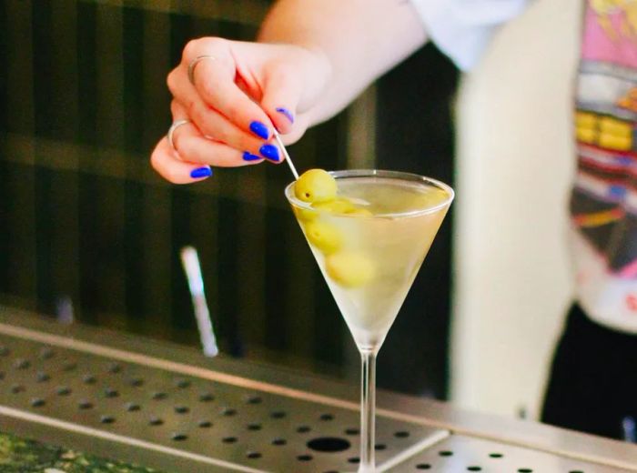 A bartender places a skewer of olives into a martini glass.