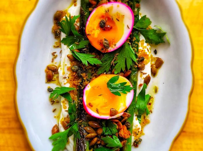 A lengthy plate of asparagus adorned with pink-dyed egg halves and a variety of herbs and garnishes.