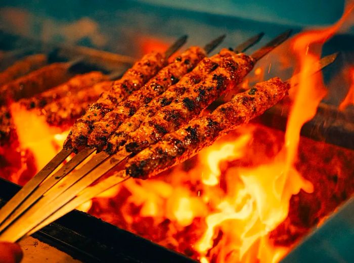 A hand holds skewers of meat above an open flame grill.