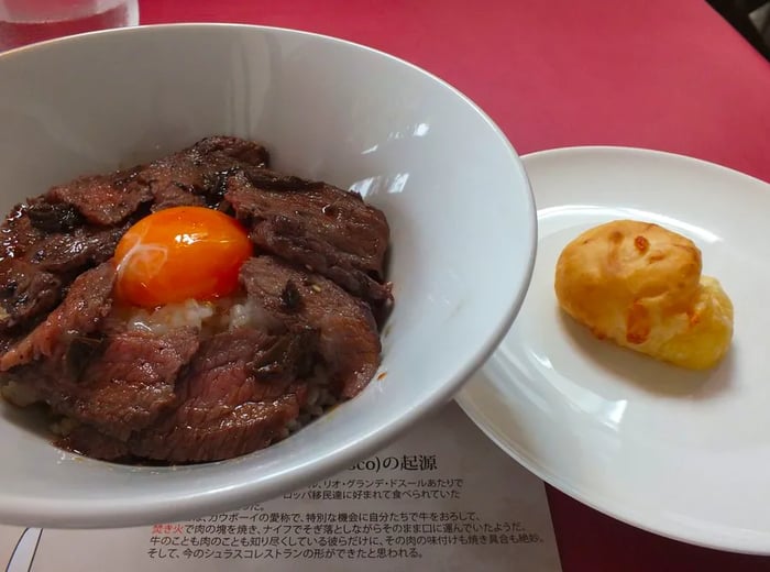 A bowl of rice adorned with slices of beef and a raw egg yolk, accompanied by a plate holding a single pan de quejo.