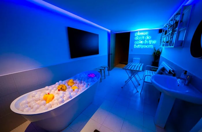 A restroom illuminated by a neon blue sign that says, “please don’t do coke in the bathroom.” It also features a bathtub filled with inflatable balls and rubber ducks.