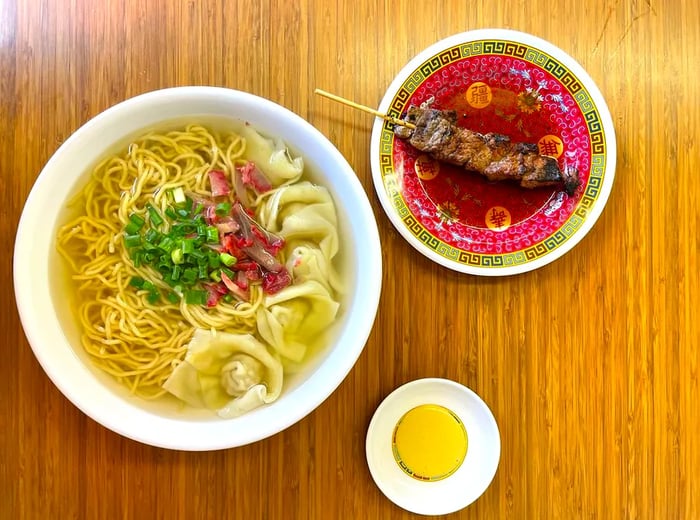 A bowl filled with noodles and dumplings, paired with a skewer of meat.