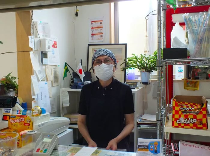 A masked staff member with a head wrap stands behind the restaurant counter, with the flags of Japan and Brazil displayed above her.