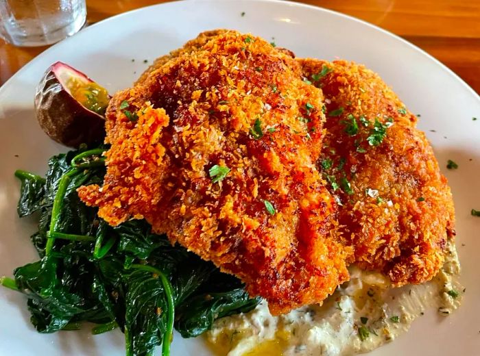 Generous portions of schnitzel served atop a bed of sautéed greens, with a slice of fresh lilikoi on the side.