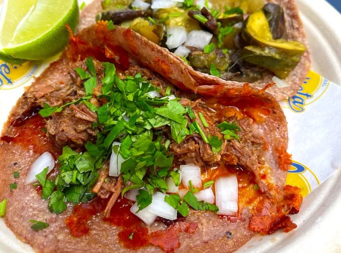 A close-up of a suadero taco, with another taco and lime wedges in the background.