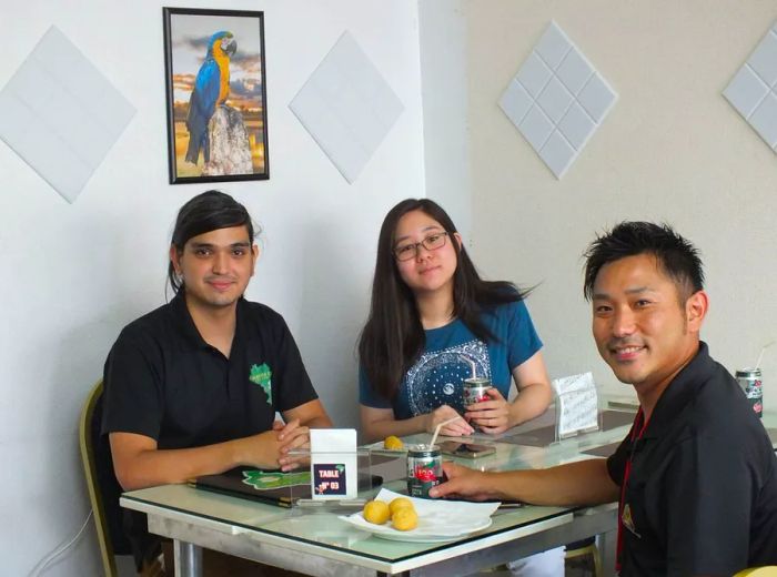 Three individuals are seated at a restaurant table, looking towards the camera.