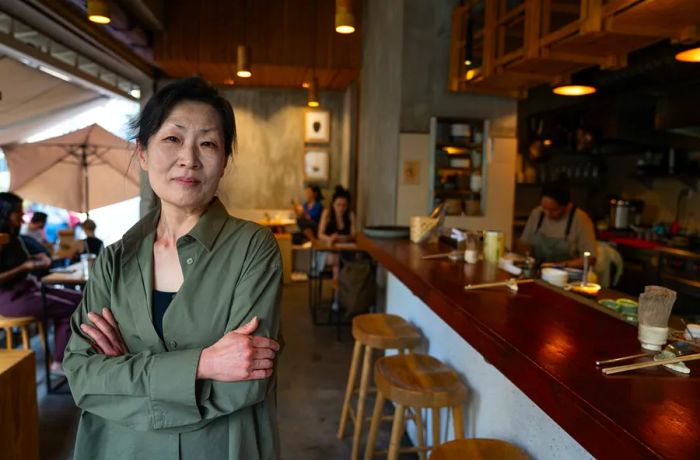 A woman stands with her arms crossed in front of a dining area and an open kitchen.