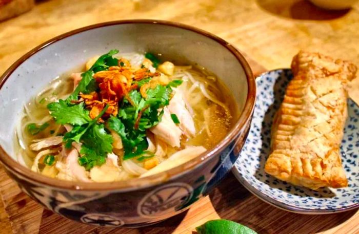 A beautifully arranged bowl of noodle soup garnished with chicken, cilantro, and various toppings, accompanied by a plate of large stuffed puff pastry.