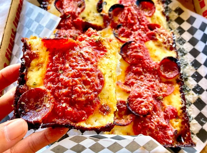 A square slice of pepperoni pizza is being held up for a photo, with the rest of the pizza visible in the background.