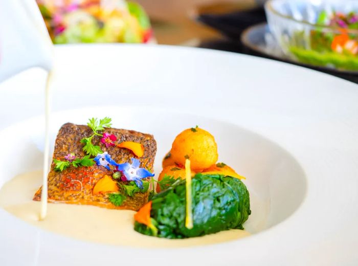 A server elegantly pours sauce over a plate featuring a fillet of kampachi alongside a vegetable side.