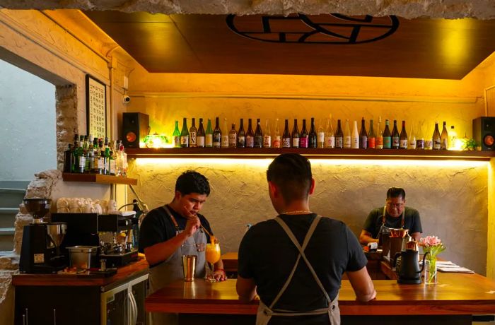 Bartenders serve drinks at a bar flanked by rustic stone walls and illuminated bottles on a back-lit shelf.