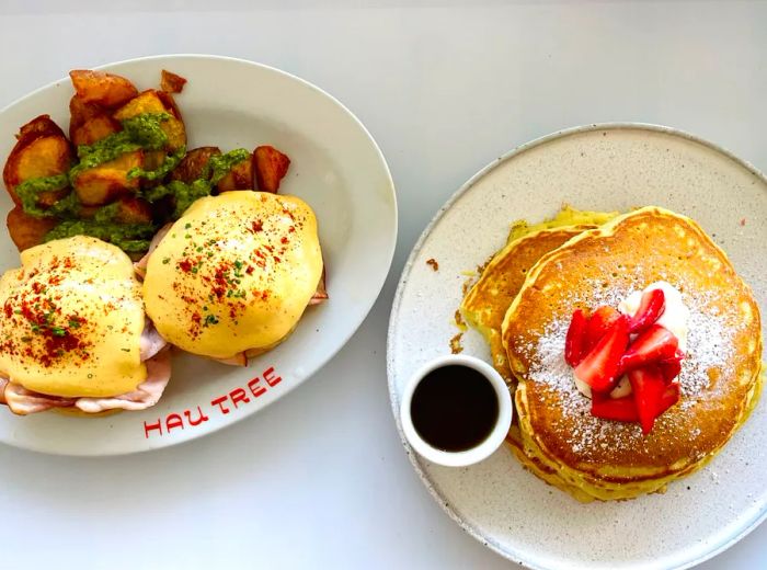 A dish of pancakes adorned with fresh strawberries and served with maple syrup, alongside a plate of eggs Benedict paired with potatoes.