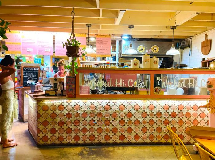 A café interior featuring bold textured wallpaper, white wooden beam ceilings, and a pastry case adorned with the café's name.