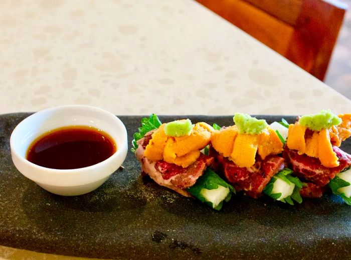 Raw beef rolls with shiso and yam, topped with uni, served on a slate with a soy sauce dipping bowl.