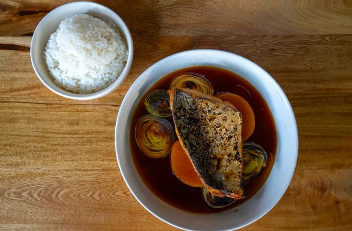 An aerial view of a large fish swimming in sauce, accompanied by spiraled vegetables and a small bowl of rice.