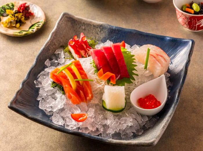 A square slate dish filled with sliced sashimi on ice, accompanied by rice, pickles, and tea.