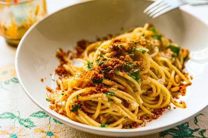 A homemade Italian pasta dish called 'pasta con le sarde,' featuring spaghetti, sardines, parsley, pine nuts, and breadcrumbs, presented on a tablecloth. It's a classic regional delicacy.