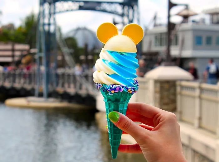 A vibrant teal ice cream cone topped with swirls of white and blue soft serve, adorned with a sprinkle-covered rim and cookies shaped like Mickey Mouse ears on top.