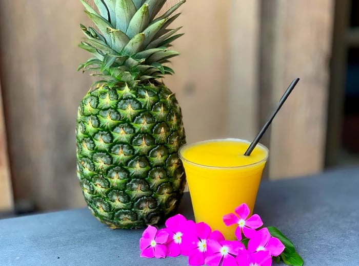 A vibrant yellow slushie in a plastic cup alongside a pineapple and a vivid purple lei.