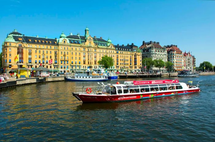 A red sightseeing boat glides past the Grand Hotel in Nybroviken, Stockholm, Sweden.