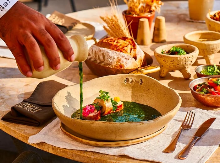 A server drizzles green sauce over a meat dish in a clay bowl, surrounded by an array of other dishes on the table.