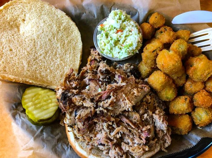 A plate featuring a chopped pork barbecue sandwich accompanied by coleslaw, pickles, and fried okra