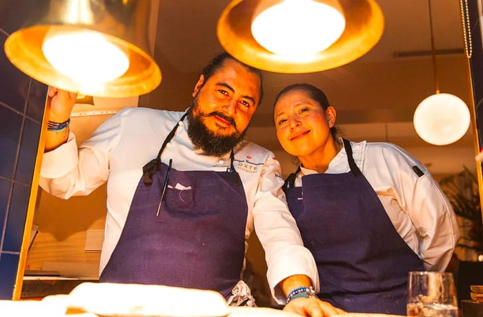 Two chefs gaze down at the camera, positioned at the height of dishes on a kitchen counter.