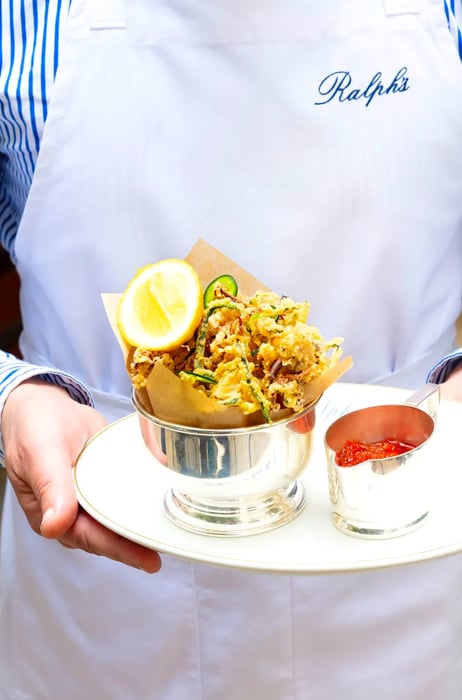 A server wearing a branded apron delivers a plate of fried squid fritters alongside sauce, both presented in silver serving ware.